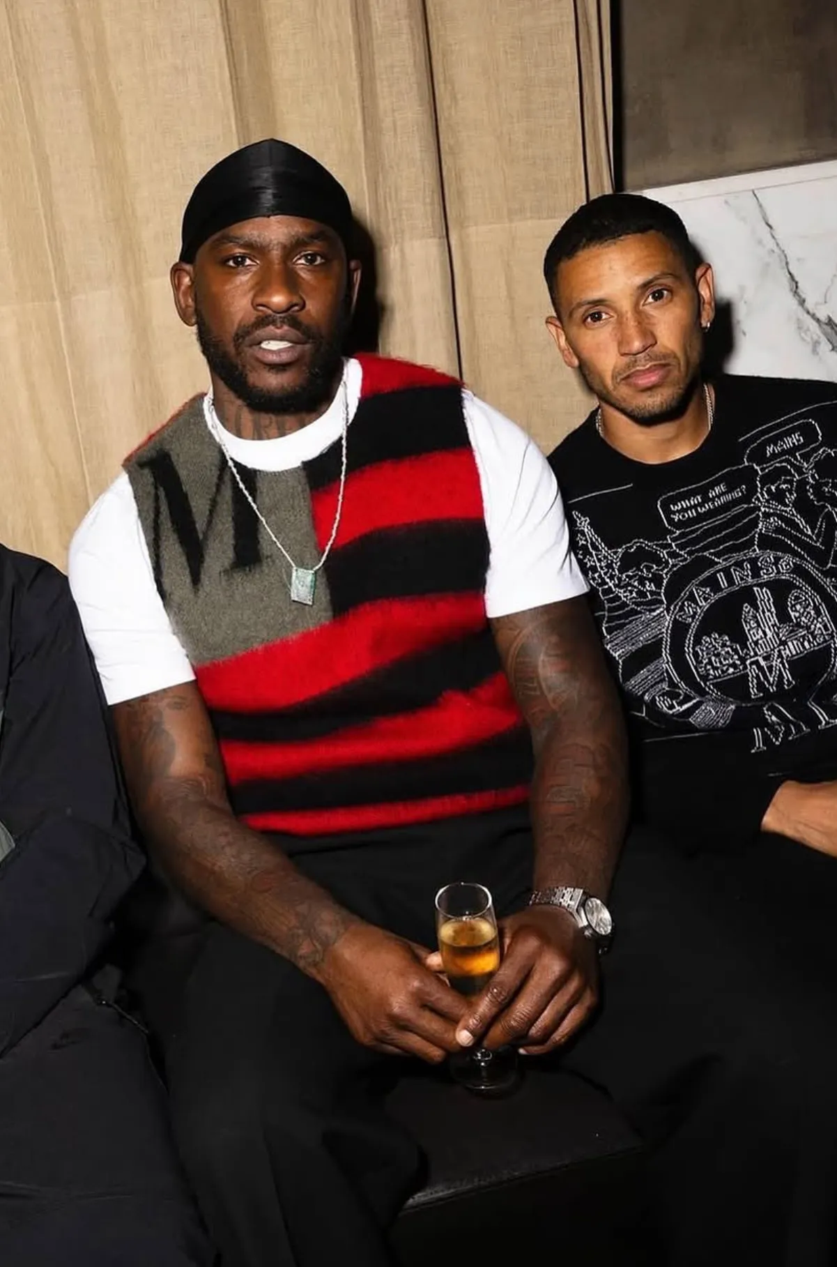 Two men at an event. One in a red and black stripes mohair vest with an M. The other in a black and white jacquard sweater.
