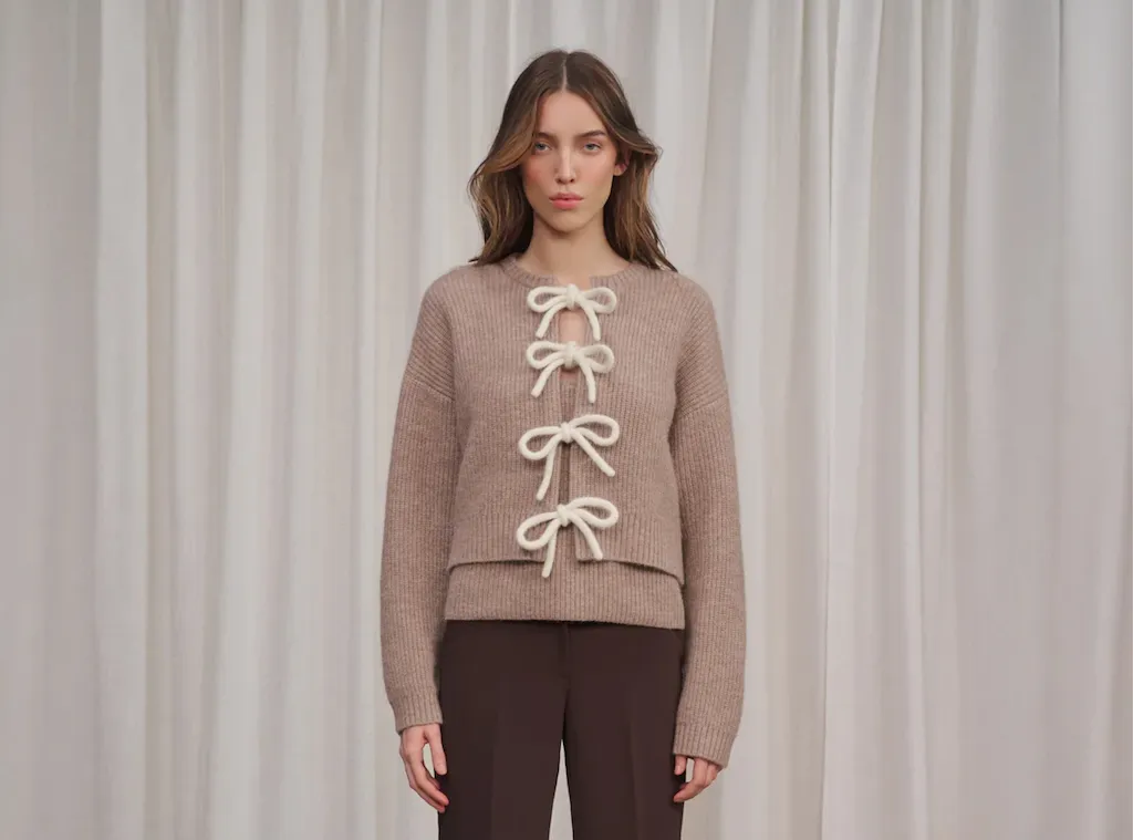 A woman in a brown knit sweater with white bows tied down the front stands against a white curtain.