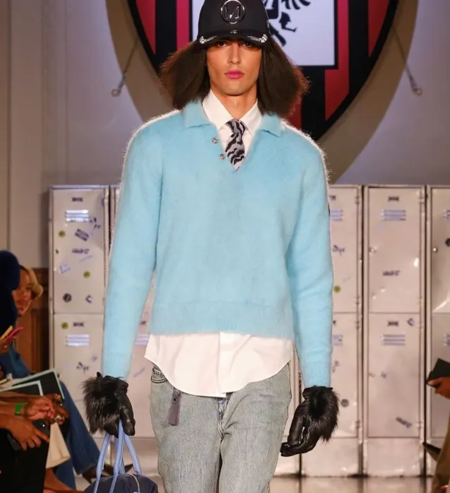 A model with long hair in a black hat, light blue mohair polo sweater, white shirt, patterned tie and jeans walks the runway.