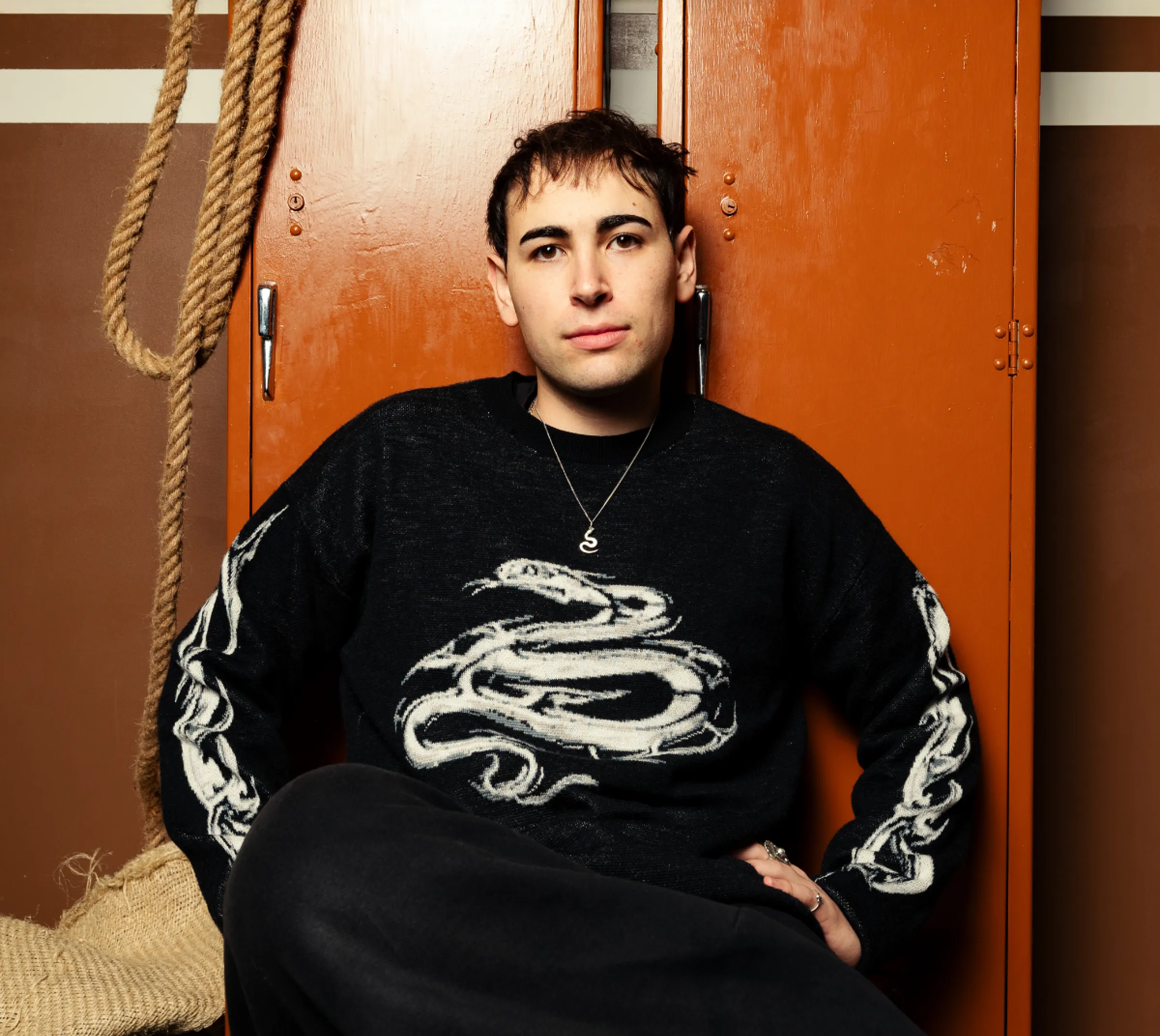 A man in a black graphic jacquard sweater with a white snake design sits, looking at the camera against storage lockers.