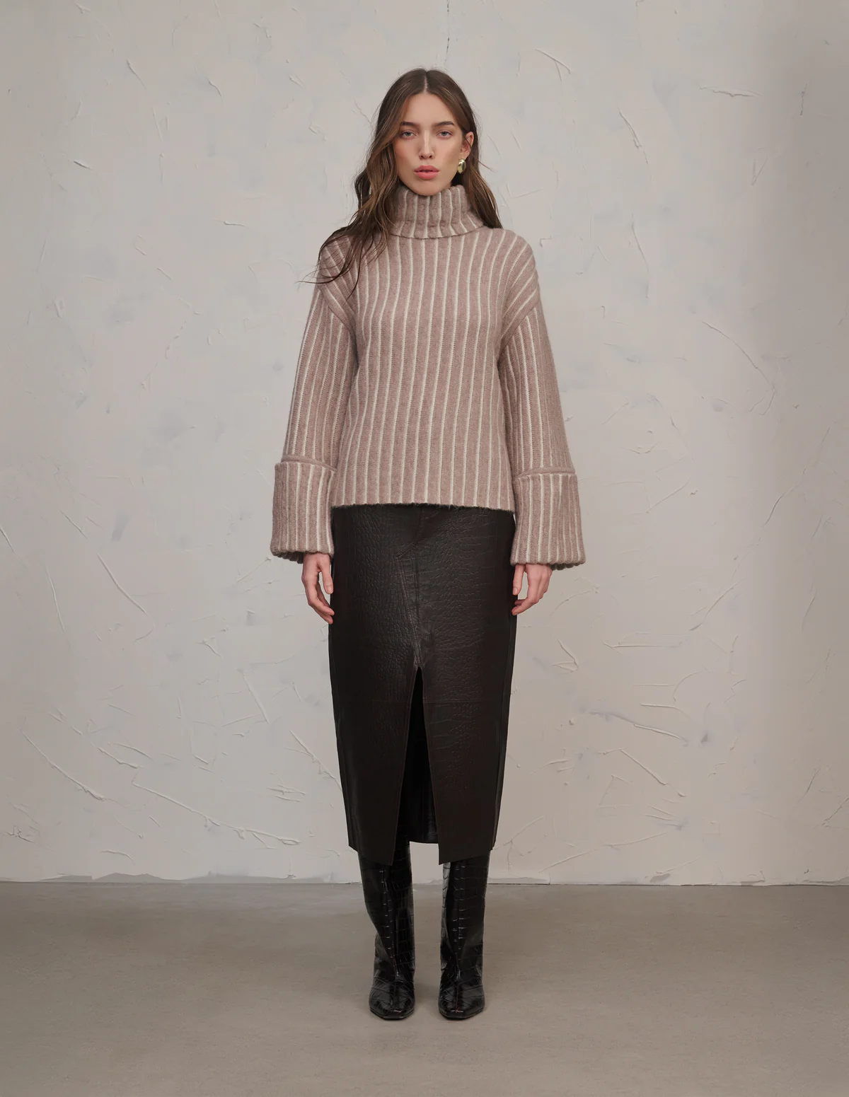 A woman in a brown striped turtleneck sweater and dark skirt stands against a textured wall.