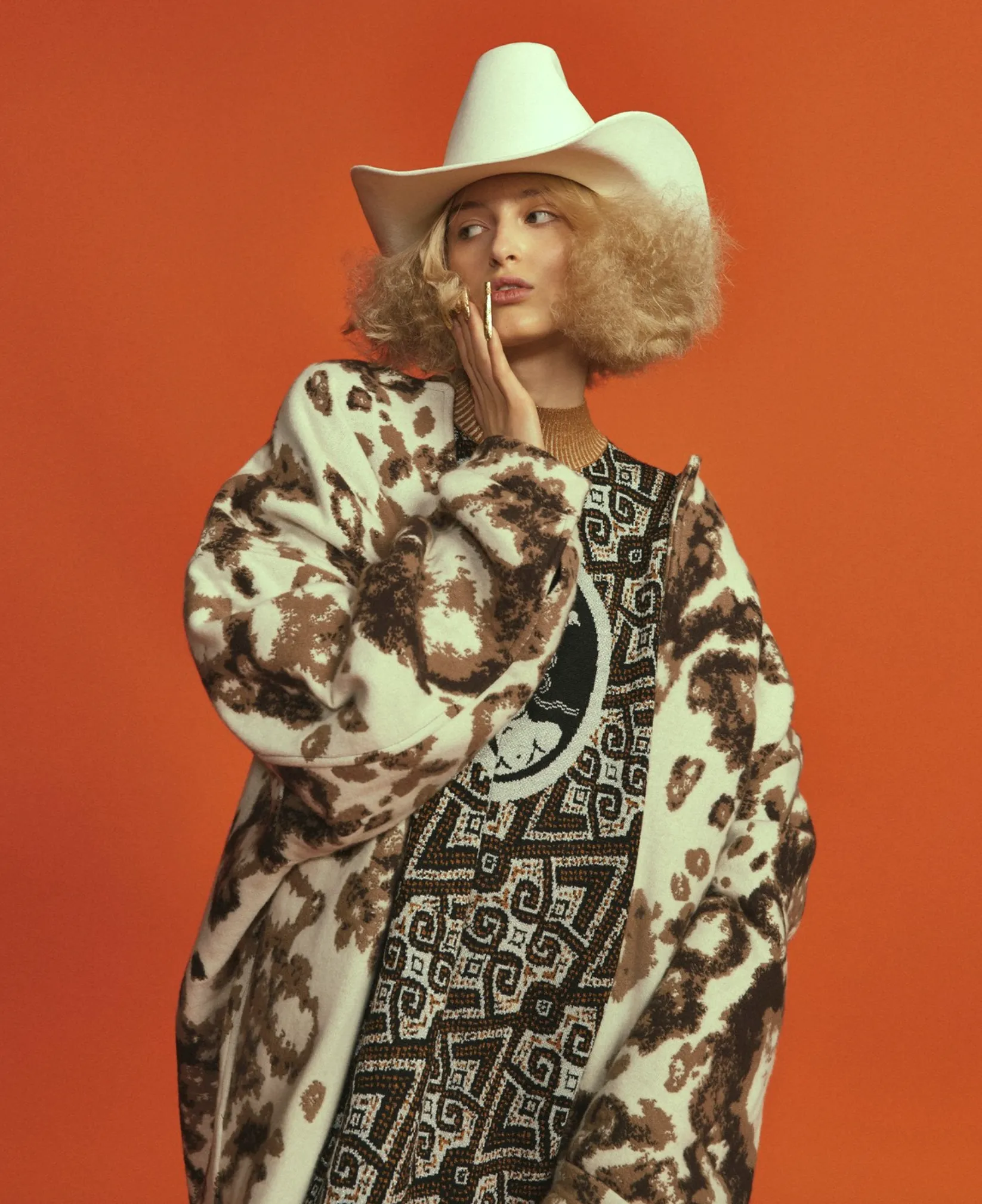 A person in a white cowboy hat, geometric jacquard knit sweater and jacket, poses against an orange background.