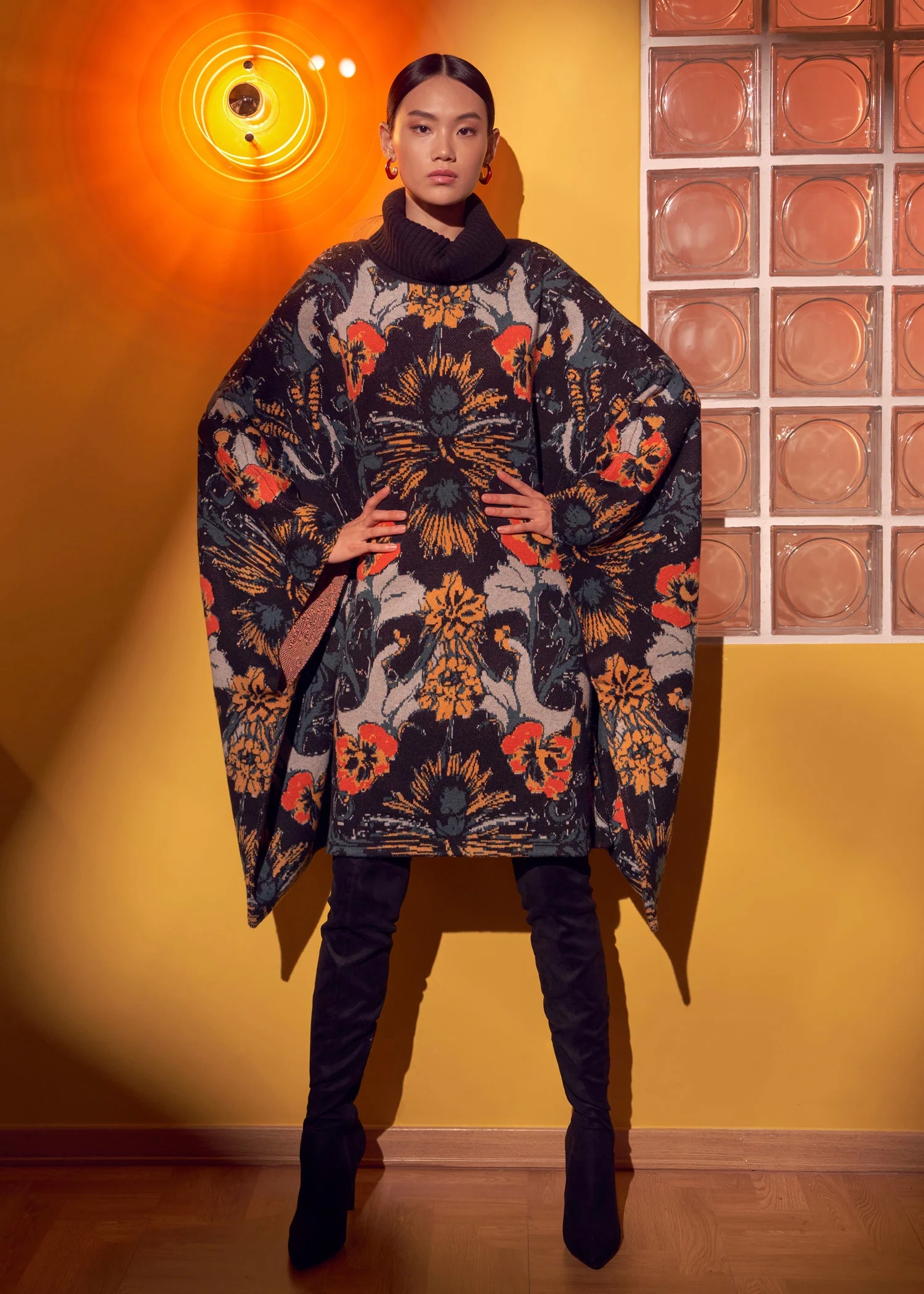 A model in a oversized, dark, floral-patterned poncho and black over-the-knee boots stands against a yellow wall.