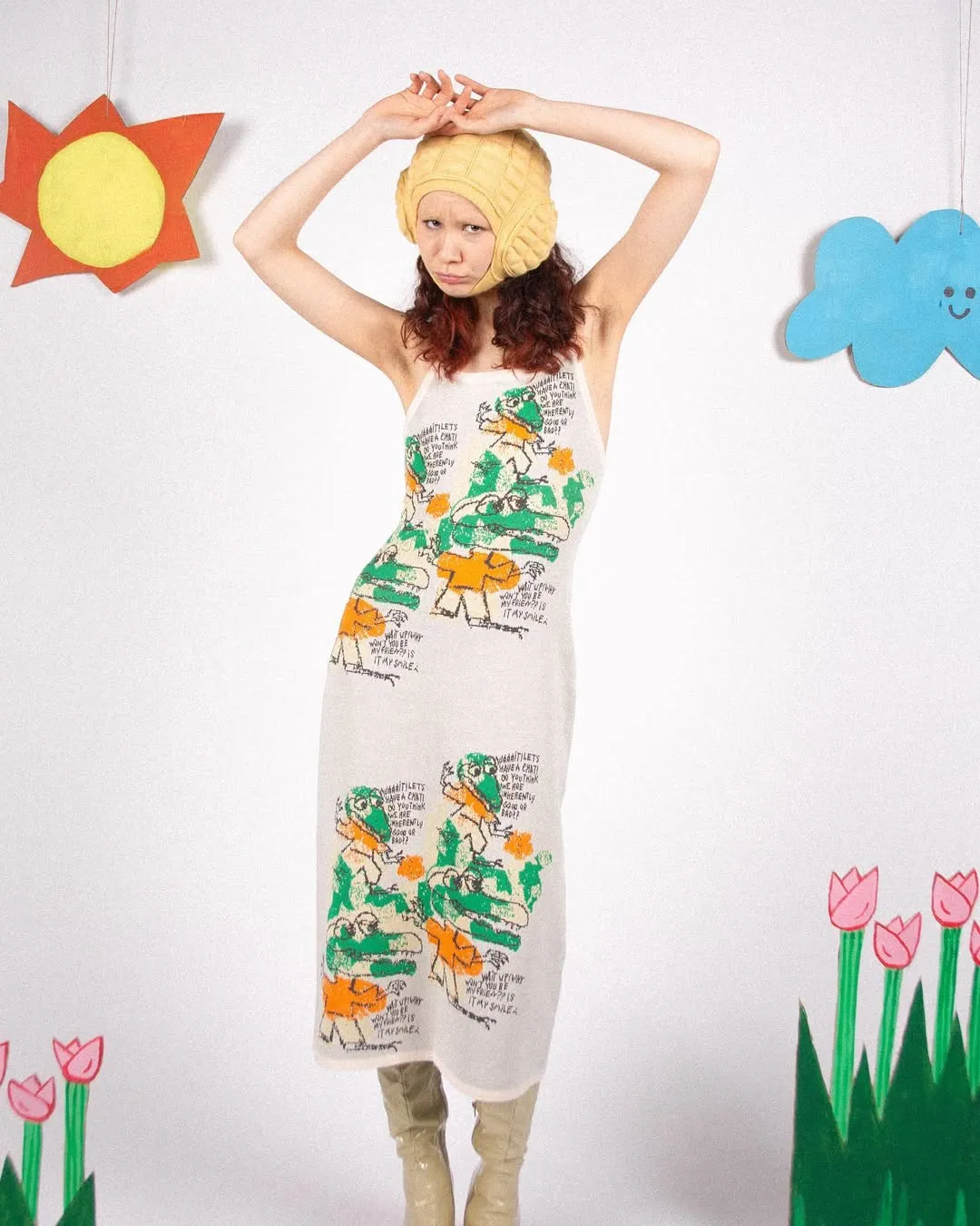 A model in a graphic jacquard knitted slip dress and yellow hat poses with arms raised, with cutout decorations in the background.