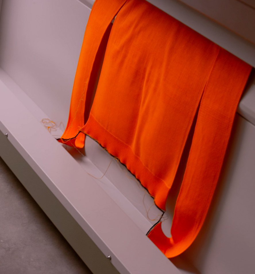 An orange jersey sweater being knitted in a knitting machine.
