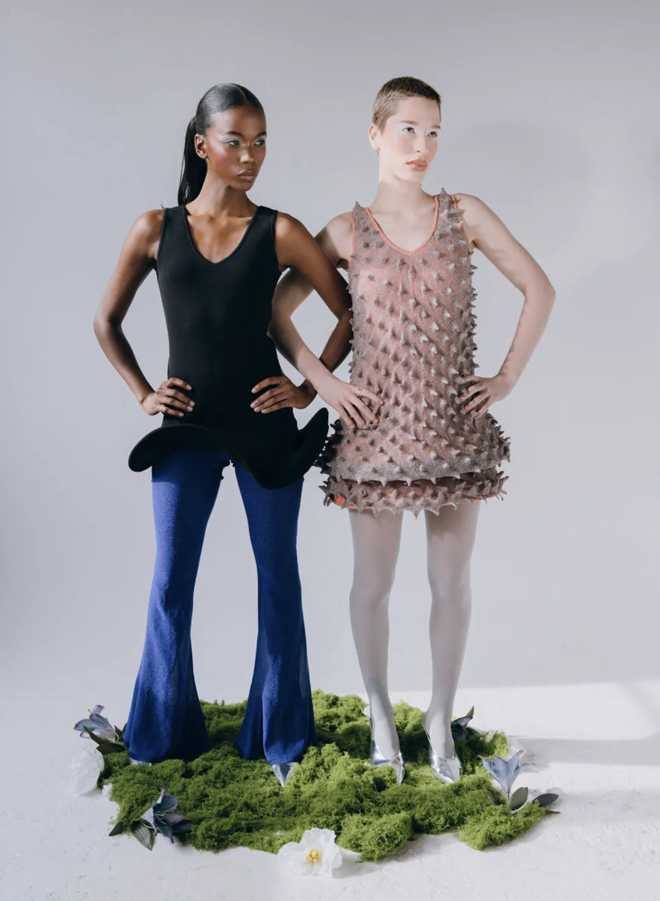 Two models pose on a mossy patch. One in black top and blue pants, the other in a spiky knitted pink dress and white tights.