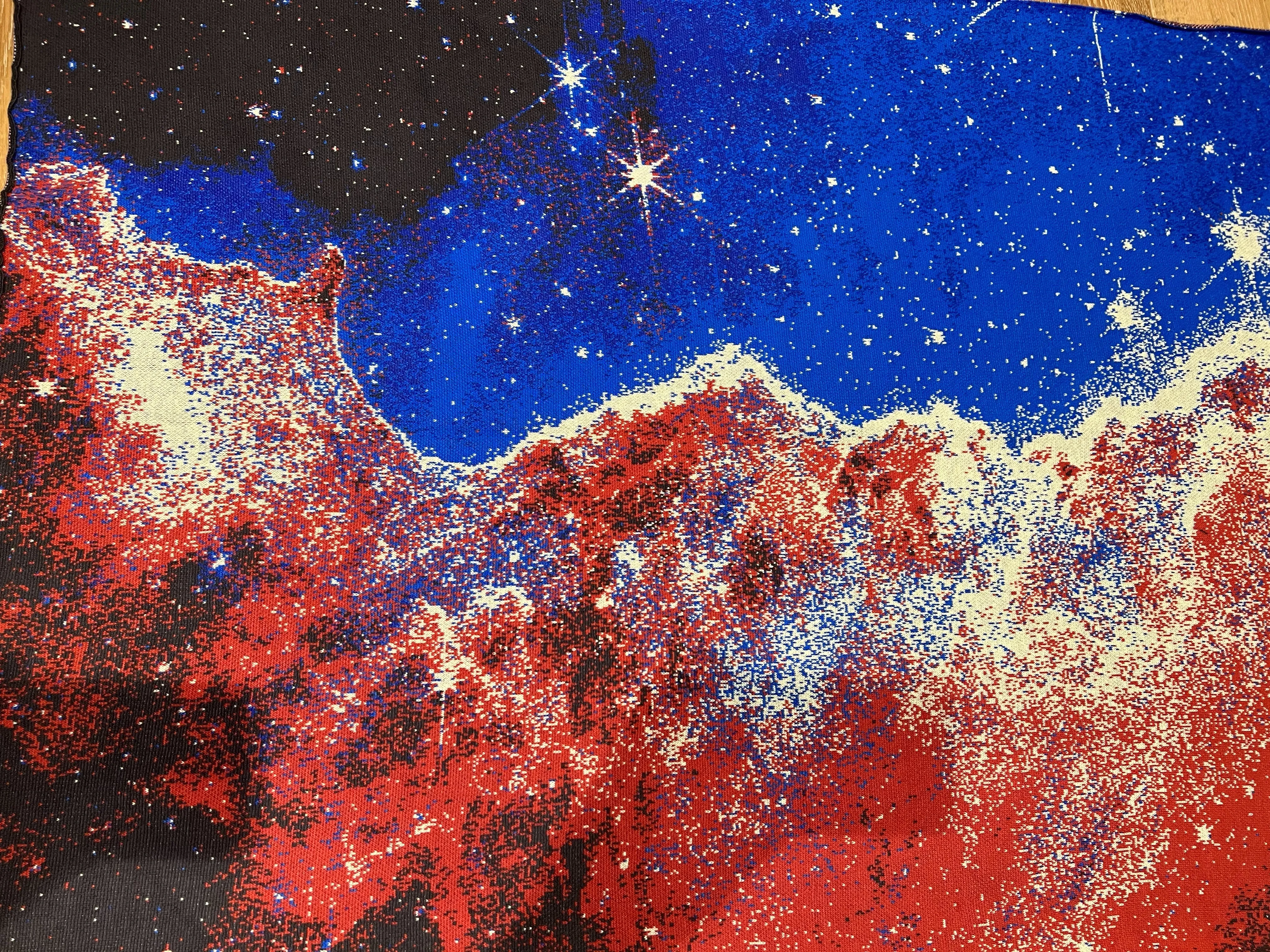 A close-up of a knitted jacquard blanket with a vibrant galaxy design featuring red, blue, white nebulae and stars.