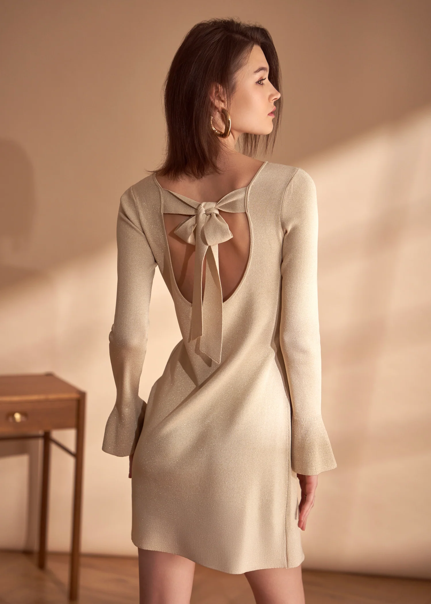 A woman in a beige knit dress with a back cut-out and bow detail, looking over her shoulder.