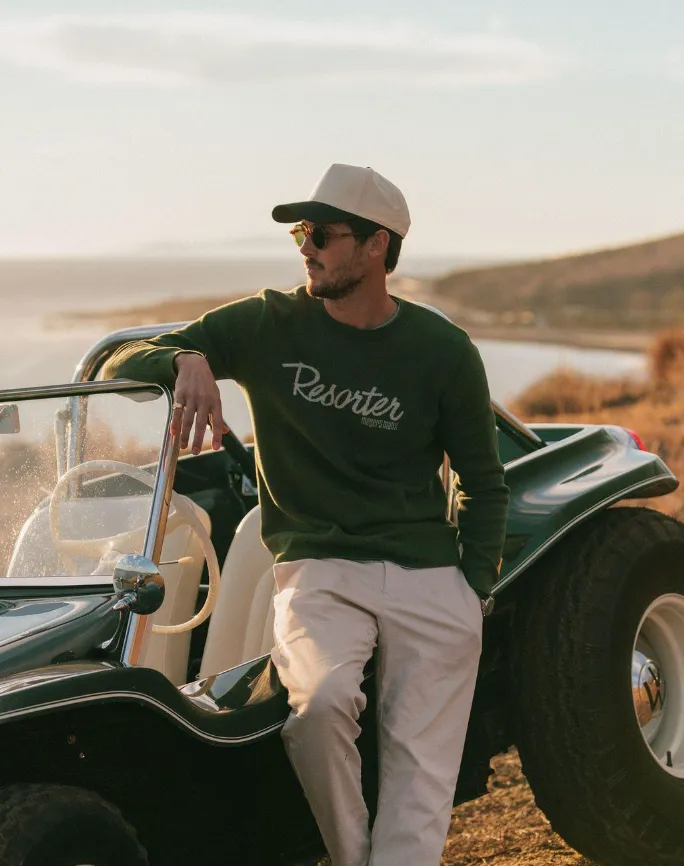 A man in a green "Resorter" knitted jacquard sweater, khaki pants, and baseball cap leans on a green dune buggy.