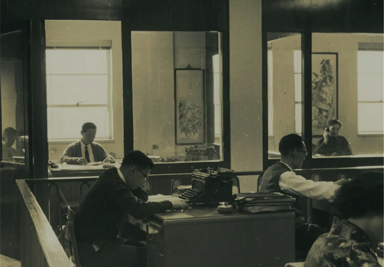 A photo of an office with people working at desks, some visible through glass partitions.