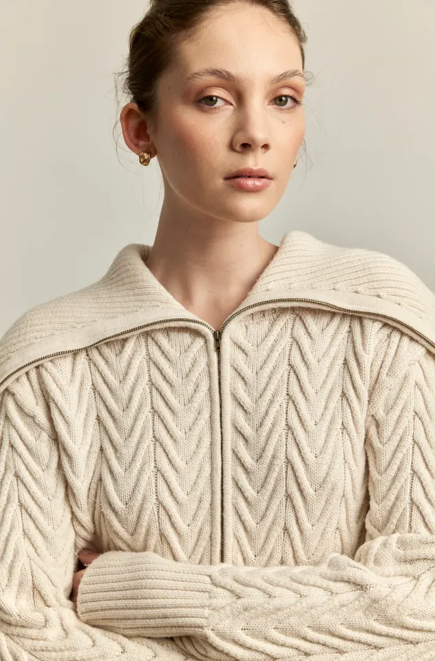 A close-up of a model wearing a cream-colored knitted chunky textured zip-up sweater.