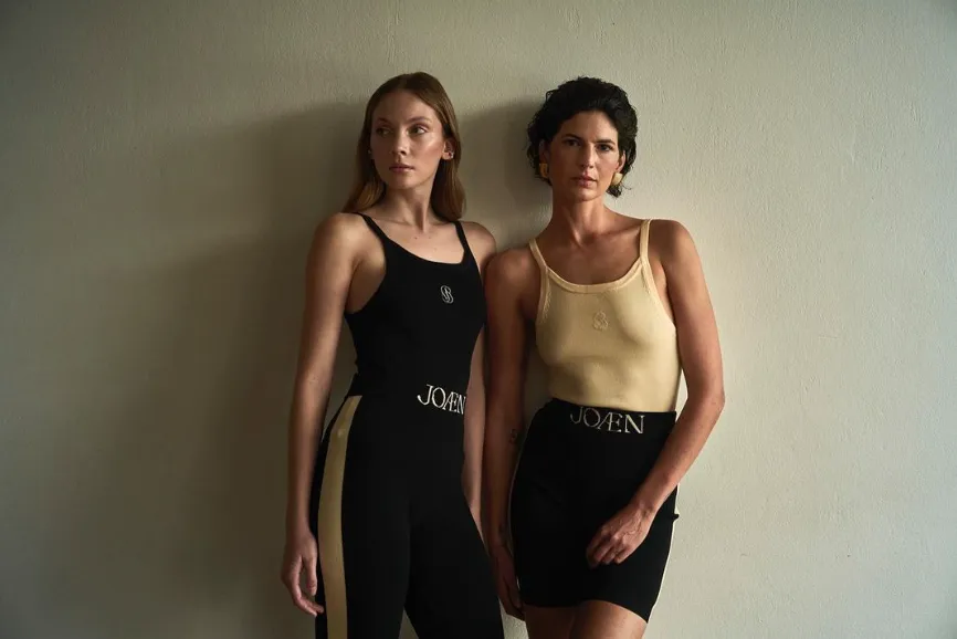Two women in knitted athleticwear. One in a black tank top and ribbed leggings, the other in a cream tank top and ribbed shorts.