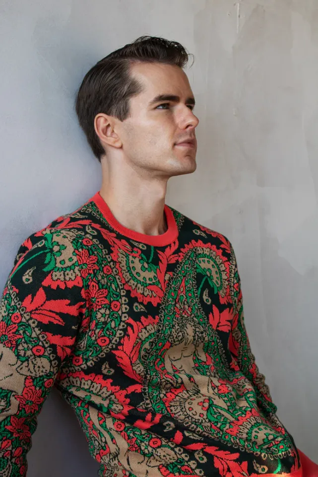 A model leaning against a wall, wearing an all-over graphic jacquard sweater in pinks, greens and blacks