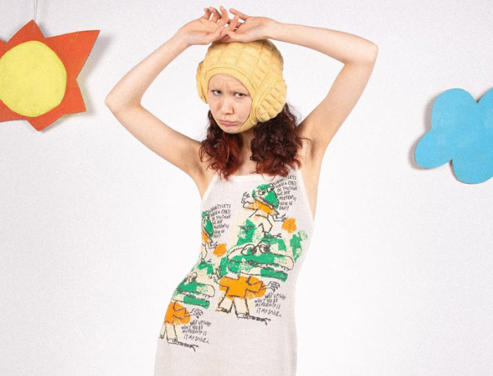A model in a graphic jacquard knitted slip dress and yellow hat poses with arms raised, with cutout decorations in the background.