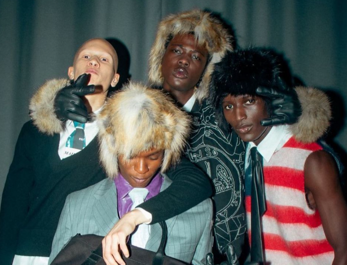 Four models in diverse outfits, including fur hats, ties, knitted mohair sweaters and vest pose together, some looking at the camera.
